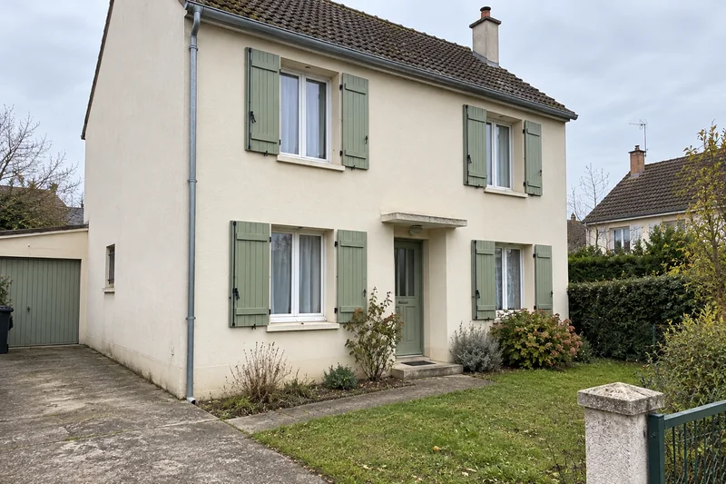 Une maison avec façade claire et volets verts, vue depuis son allée